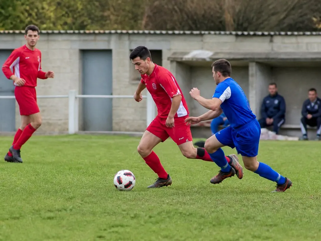 Match de football amateur sur gazon : un joueur en bleu presse un joueur en rouge devant les vestiaires du stade